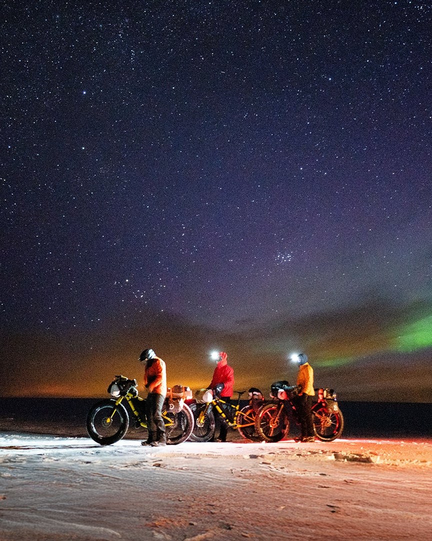 Syklister med fatbikes og hodelykter under nordlyset på en snødekt slette