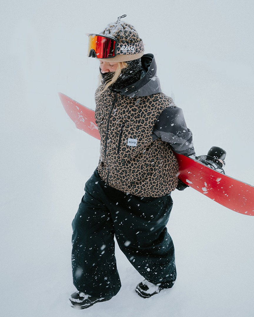 Snowboarder i Eivy-jakke og -lue bærer brettet gjennom snøværet